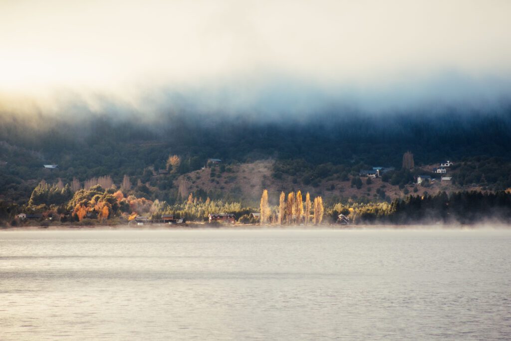 Lago Meliquina cubierto de niebla al atardecer, con álamos en otoño y casas de madera asomando entre la vegetación de la costa