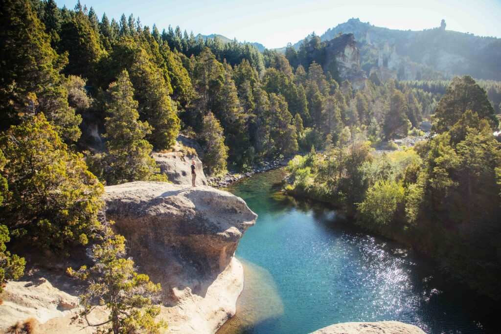 Ruta los Siete Lagos. Una persona contemplando un pozón de agua turquesa desde una roca, rodeada de cipreses y formaciones rocosas en la cordillera neuquina
