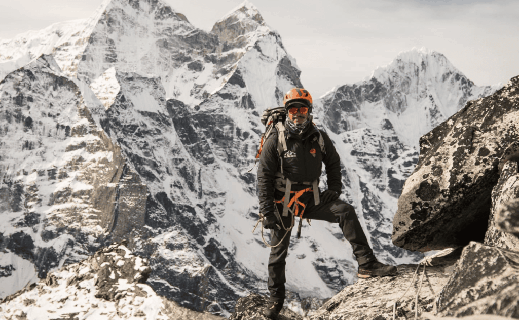 Montañista con casco y arnés de escalada posa sobre una cresta rocosa con picos nevados del Himalaya de fondo