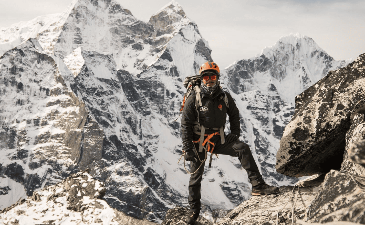 Montañista con casco y arnés de escalada posa sobre una cresta rocosa con picos nevados del Himalaya de fondo