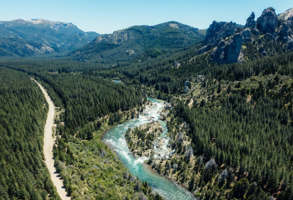 Vista aérea de un camino de ripio bordeando un río turquesa entre bosque de pinos, con formaciones rocosas de fondo, fotografía de Gustavo Arias