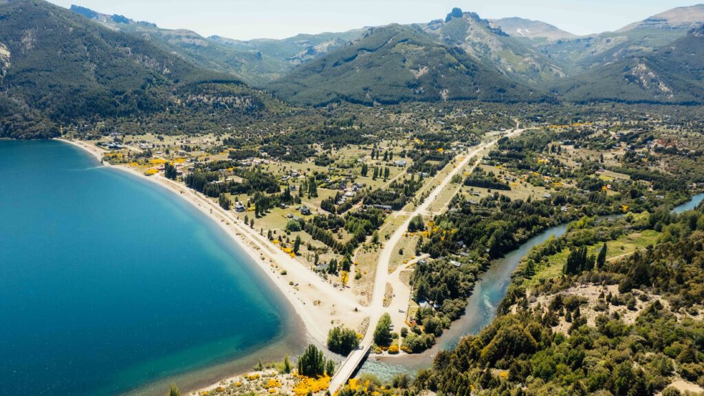 ¿Cómo llegar a Villa Meliquina? Vista aérea de un pueblo patagónico a la orilla de un lago, con playa, calles arboladas y la desembocadura de un río