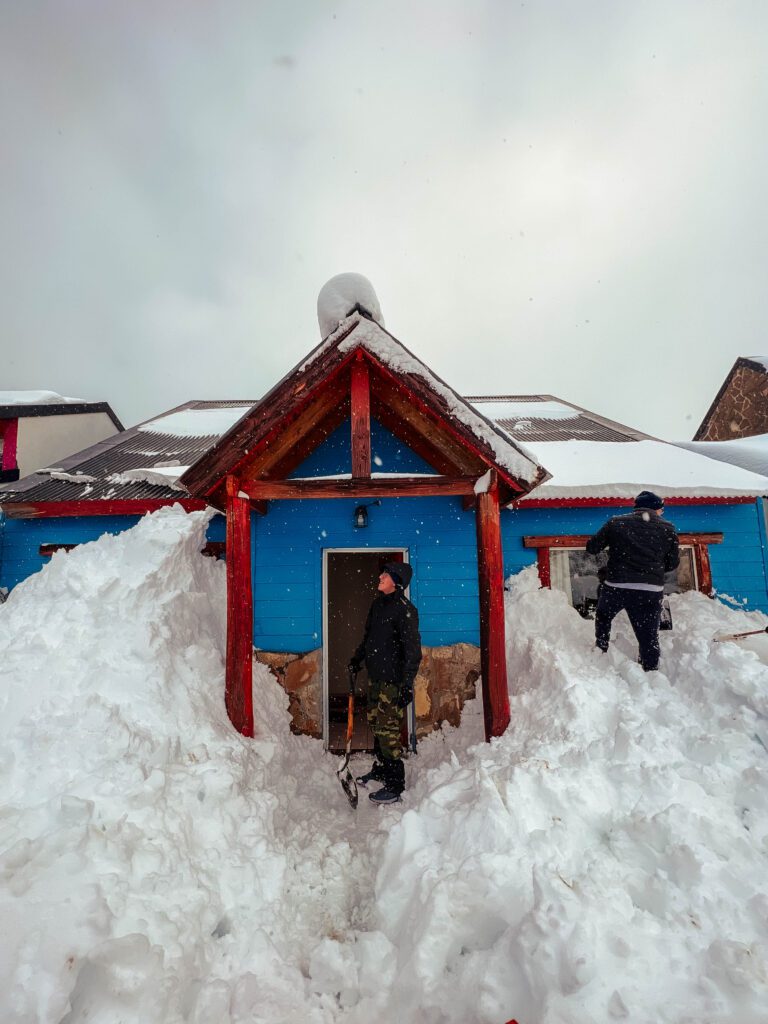 caviahue en invierno frente de una casa completamente nevada con dos personas sacando la nieve con palas
