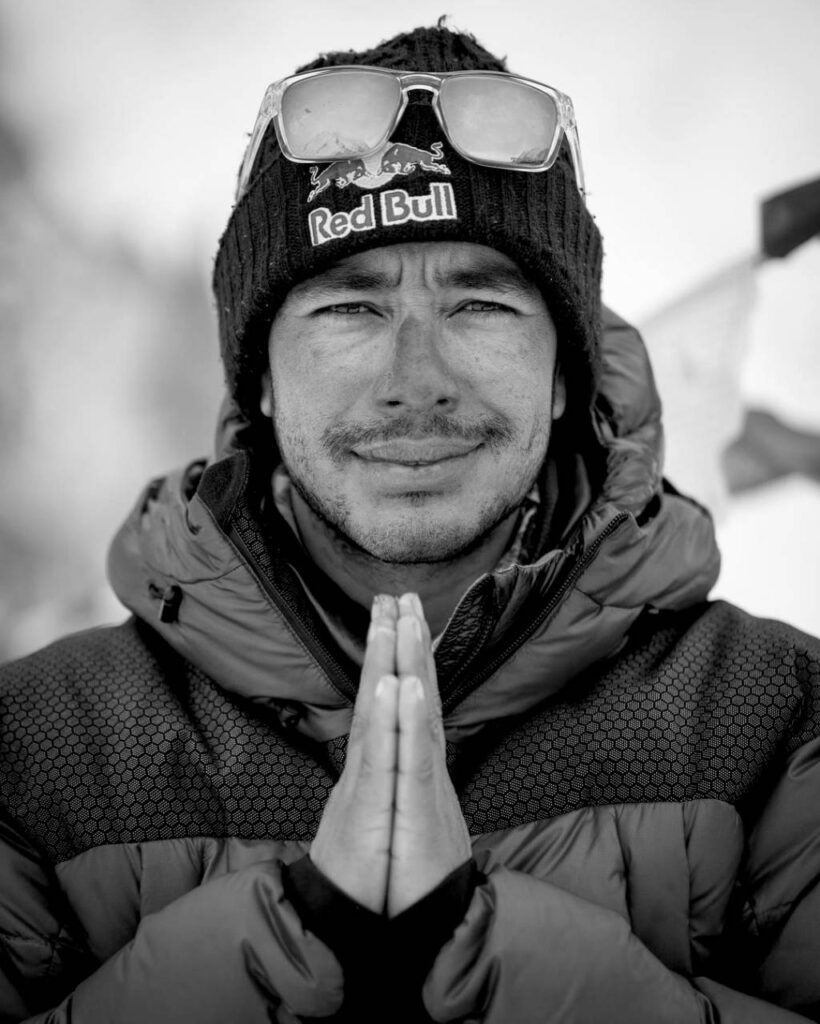 Retrato en blanco y negro de Nirmal Purja con gorro Red Bull y anteojos de sol sobre la cabeza, sonriendo con las manos juntas en gesto de agradecimiento, vestido con campera de montaña